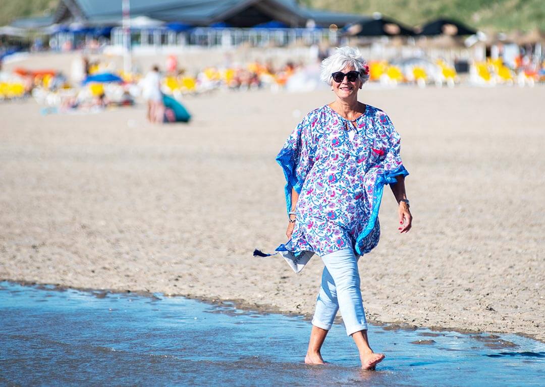 Mevrouw loopt op het strand in blauwe kaftan en donkere zonnebril